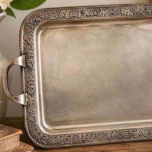 May include: A rectangular silver-toned metal serving tray featuring a raised border with an intricate embossed floral and grape vine pattern. The tray includes two curved side handles and a smooth, slightly worn central surface area.
