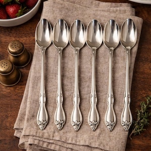 May include: Six silver spoons with ornate handles are arranged on a tan linen napkin. The spoons are placed on a dark wooden surface, with a bowl of strawberries and brass salt and pepper shakers in the background. A white plate with flowers is also visible.