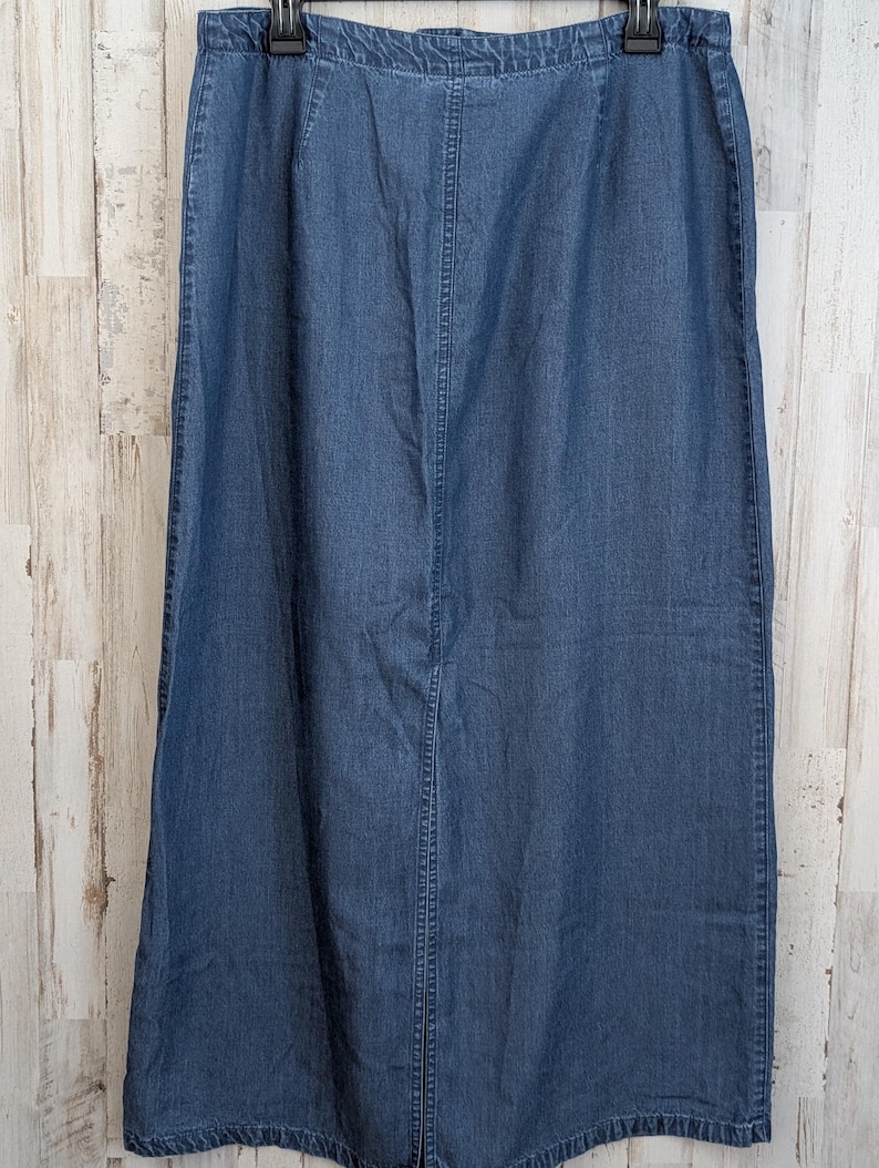 May include: A long, medium-wash blue denim skirt featuring a straight silhouette, a high-waisted design with an elasticated waistband, and a prominent vertical centre seam running down the front, finished with a simple hem at the bottom edge.
