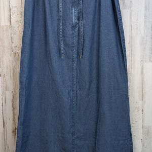 May include: A long, blue denim skirt featuring an elasticated waistband with a matching fabric drawstring tie. The skirt has a straight silhouette, a central vertical seam running down the front, and a hem that falls below the knee.