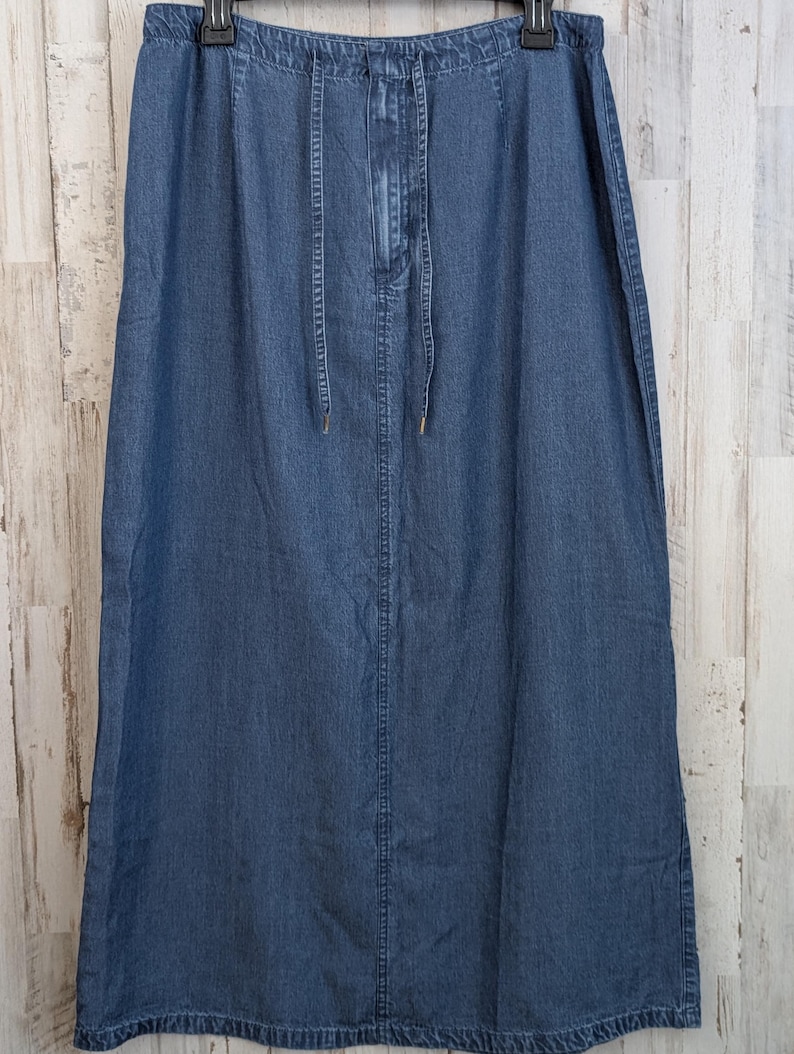 May include: A long, blue denim skirt featuring an elasticated waistband with a matching fabric drawstring tie. The skirt has a straight silhouette, a central vertical seam running down the front, and a hem that falls below the knee.