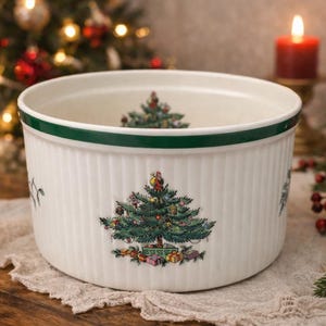 May include: A white ceramic bowl with vertical ribbed sides and a green rim. The exterior features a colorful illustration of a decorated Christmas tree with wrapped gifts at the base. A smaller matching tree design is visible on the interior bottom.