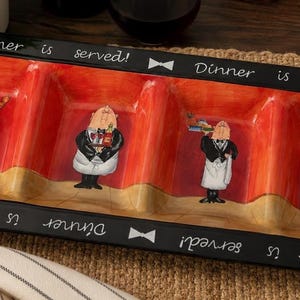 May include: A rectangular ceramic serving tray with four divided sections, featuring a vibrant red interior and a black border. The black rim displays the repeated white text "Dinner is served!" and small white bow tie icons. Each section contains a painted illustration of a waiter in a tuxedo and apron.