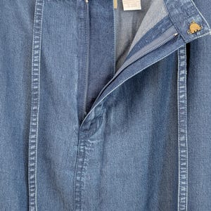 May include: A blue denim skirt featuring a front zip closure, a waistband with a metal button, and two vertical fabric strips running down the front. A white interior label displays garment information, and the fabric has a light wash finish.