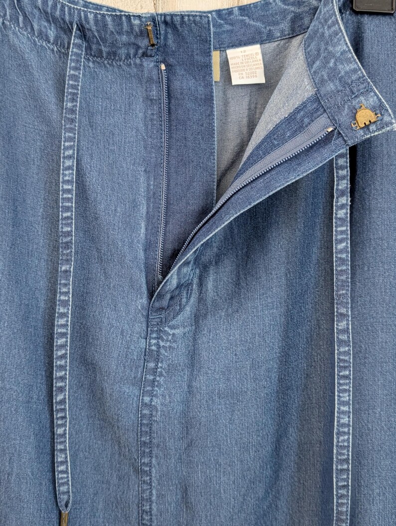 May include: A blue denim skirt featuring a front zip closure, a waistband with a metal button, and two vertical fabric strips running down the front. A white interior label displays garment information, and the fabric has a light wash finish.