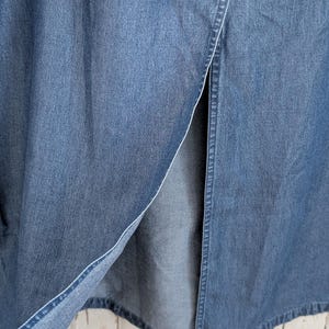 May include: A long blue denim skirt featuring a prominent vertical front slit. The fabric displays a classic medium-wash indigo texture with visible stitching along the edges of the opening, showcasing the durable cotton material and construction.