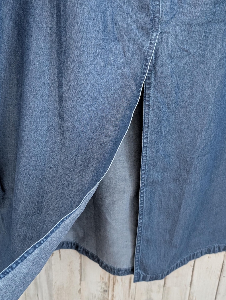 May include: A long blue denim skirt featuring a prominent vertical front slit. The fabric displays a classic medium-wash indigo texture with visible stitching along the edges of the opening, showcasing the durable cotton material and construction.