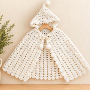 May include: A cream-colored crocheted hooded cape featuring a classic granny square pattern, a pointed hood topped with a small pom-pom, and a front tie closure finished with two matching pom-poms. The edges have a scalloped crochet trim.