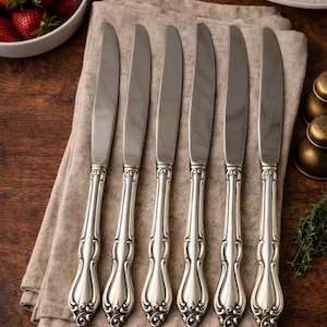 May include: A set of six silver-toned dinner knives featuring smooth, polished blades and ornate, embossed handles with intricate scrollwork detailing. The knives are arranged in a parallel row, showcasing a consistent metallic finish and design.