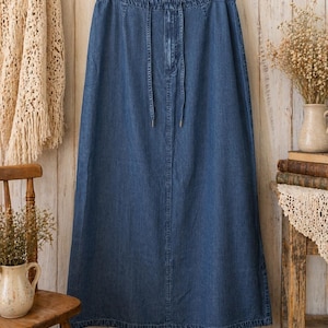 May include: A long, medium-wash blue denim skirt featuring an elasticated waistband with a functional drawstring tie. The skirt has a straight, floor-length silhouette with visible vertical seam detailing down the centre front and a hemmed bottom edge.