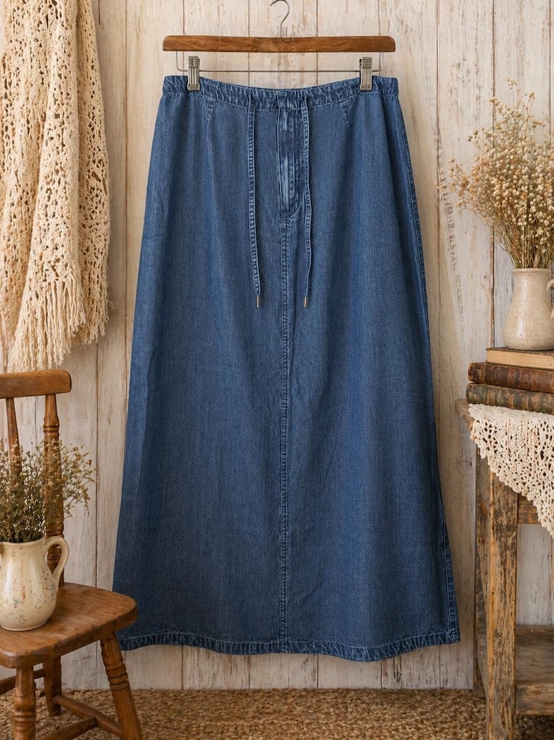 May include: A long, medium-wash blue denim skirt featuring an elasticated waistband with a functional drawstring tie. The skirt has a straight, floor-length silhouette with visible vertical seam detailing down the centre front and a hemmed bottom edge.