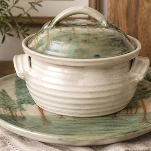 May include: A ceramic lidded pot with horizontal ridges and two side handles, resting on a matching round plate. Both pieces feature a hand-painted landscape design with green trees and brown trunks against a cream-colored glazed background.