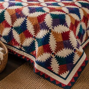 May include: A quilt featuring a repeating pineapple block pattern in shades of red, blue, purple, and gold, contrasted with off-white fabric. The textile displays intricate geometric stitching throughout, finished with a decorative border.