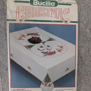 Puede incluir: Un kit de mantel de punto de cruz navide&ntilde;o Bucilla. El paquete muestra un mantel blanco con un borde festoneado rojo, con dise&ntilde;os de &aacute;rboles de Navidad y renos. El texto "CHRISTMAS" se muestra en rojo y verde.