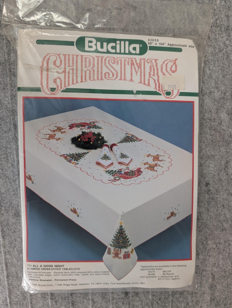 Puede incluir: Un kit de mantel de punto de cruz navide&ntilde;o Bucilla. El paquete muestra un mantel blanco con un borde festoneado rojo, con dise&ntilde;os de &aacute;rboles de Navidad y renos. El texto "CHRISTMAS" se muestra en rojo y verde.