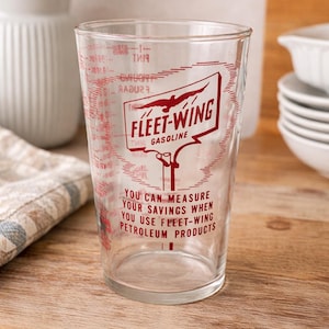May include: Clear glass measuring cup with red markings and the "Fleet-Wing Gasoline" logo. The text on the cup reads "You can measure your savings when you use Fleet-Wing Petroleum Products."
