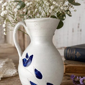 May include: A white ceramic pitcher with a handle, featuring a blue floral design. The pitcher is filled with white flowers and greenery. The pitcher is sitting on a wooden surface.