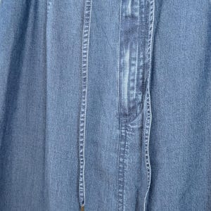 May include: A blue denim skirt featuring an elasticated waistband with a drawstring closure. The fabric has a medium wash finish with vertical stitching details along the front panel. Two long, thin denim drawstrings hang down from the centre.