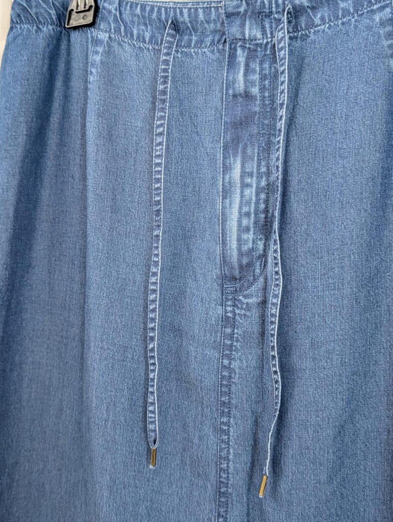 May include: A blue denim skirt featuring an elasticated waistband with a drawstring closure. The fabric has a medium wash finish with vertical stitching details along the front panel. Two long, thin denim drawstrings hang down from the centre.