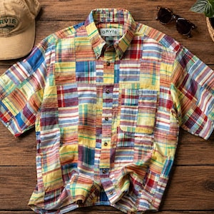May include: A short-sleeved, button-down shirt with a patchwork design in various colors, including yellow, red, blue, and green. The shirt has a pocket on the chest and the brand name "ORVIS" is visible on the collar. A tan baseball cap and sunglasses are also in the image.