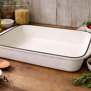 May include: A white rectangular baking dish with a black trim sits on a wooden surface. The dish is surrounded by ingredients like tomatoes, herbs, and spices. A wooden spoon and small bowls are also present.