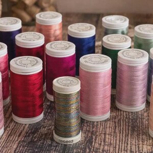 May include: A collection of colorful spools of thread in various shades, including pink, red, yellow, and blue. The spools are arranged on a wooden surface, with a neutral background. The thread is tightly wound on white spools.