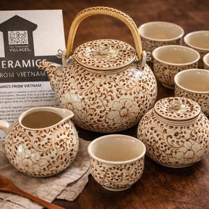 May include: Ceramic tea set from Vietnam, featuring a teapot with a woven handle, a creamer, a sugar bowl, and six teacups. The set is decorated with a brown floral pattern on a cream background. A wooden spoon and a card are also present.