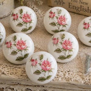 May include: Set of eight white ceramic cabinet knobs. Each knob features a pink rose design with green leaves. The knobs are round and have a glossy finish, perfect for adding a touch of floral elegance to furniture.