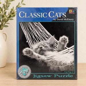May include: A rectangular jigsaw puzzle box titled Classic Cats by David McEnery. The box features a black and white photograph of a kitten sleeping in a white rope hammock. The text reads Classic Cats, Cat Nap II, and 529 pieces.