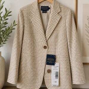 May include: A beige and white gingham blazer with a notched lapel, two brown buttons, and long sleeves. The garment features a Pendleton brand label at the inner neckline and a hanging retail tag with product details attached to the front button.