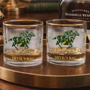 May include: Two clear whiskey glasses with gold rims and a horse racing graphic. The glasses have the text "DEVIL'S BAG" at the base. The glasses are on a gold-rimmed tray.