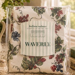 May include: A Waverly balloon valance packaged in clear plastic, featuring a cream-colored fabric with a pattern of green leaves and clusters of red and blue grapes. The label reads: balloon valance, fits window 36"-48" wide (92cm-122cm).