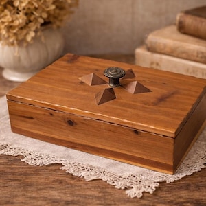 May include: A rectangular wooden box with a hinged lid. The box is made of light brown wood and features a decorative metal knob on top. The lid is adorned with carved pyramid-shaped accents. The box is placed on a lace doily.