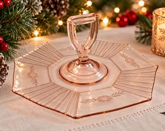 May include: A pale pink glass serving tray with a hexagonal shape and a central vertical handle. The tray features etched floral patterns on alternating panels and radiating linear textures on the remaining sections of the glass surface.