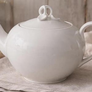 May include: A white ceramic teapot with a rounded body, curved spout, and a loop handle. The lid features a decorative floral-like knob. The teapot is sitting on a neutral-colored linen cloth.