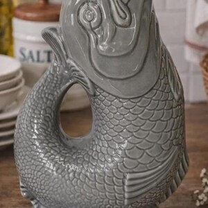 May include: A gray ceramic pitcher shaped like a fish. The pitcher has a wide opening at the top and a handle formed by the fish's tail. The body is covered in scales, and the fish's head is detailed with eyes and fins. A decorative kitchenware item.