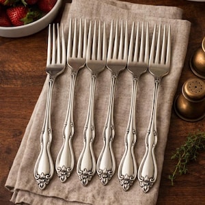 May include: A set of six silver forks with ornate handles, arranged on a beige linen napkin. The forks have a classic design with decorative detailing. The scene is set on a dark wooden surface, suggesting a dining setting.