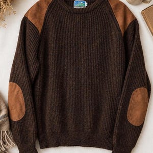 May include: A dark brown ribbed knit sweater featuring tan suede patches on the shoulders and elbows. The garment has a crew neckline and long sleeves with a label at the inner collar. The texture is thick and consistent throughout the body.