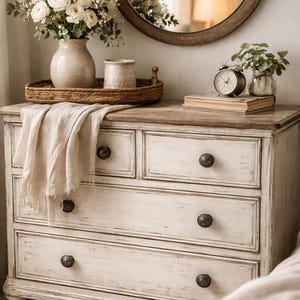 May include: A white painted wooden dresser with four drawers and dark knobs. A round mirror hangs above the dresser. A vase of white flowers, a small jar, and a woven tray sit on top. A small alarm clock and a glass vase with greenery are also on the dresser.