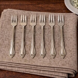 May include: A set of six silver-toned metal forks arranged in a row on a folded beige linen napkin. Each fork features a slender handle with a decorative, embossed floral motif at the base and four tines at the top, showcasing a classic design.