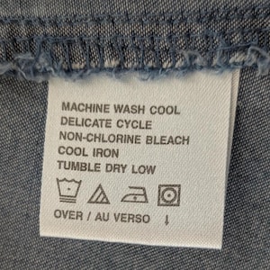 May include: A white rectangular fabric care label sewn onto blue denim material. The text reads: MACHINE WASH COOL, DELICATE CYCLE, NON-CHLORINE BLEACH, COOL IRON, TUMBLE DRY LOW, OVER / AU VERSO. Four care symbols are printed below the text.