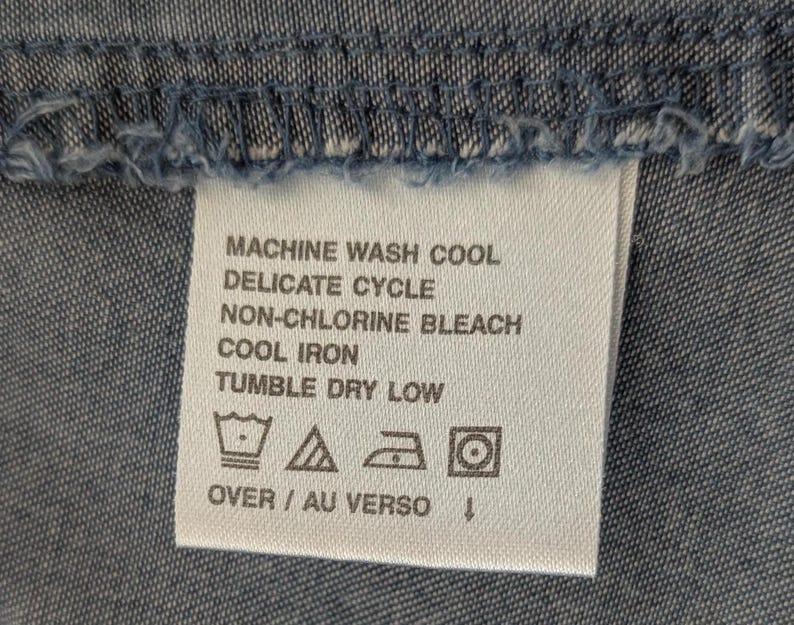 May include: A white rectangular fabric care label sewn onto blue denim material. The text reads: MACHINE WASH COOL, DELICATE CYCLE, NON-CHLORINE BLEACH, COOL IRON, TUMBLE DRY LOW, OVER / AU VERSO. Four care symbols are printed below the text.
