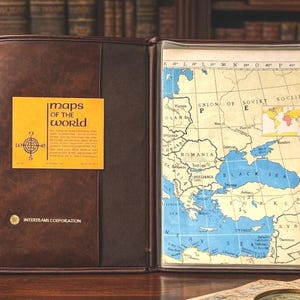 May include: Open brown leather map case with a vintage map of Europe and the Middle East. The map is framed within the case. A yellow sticker on the left side reads "maps of the world". The case is open on a wooden surface.