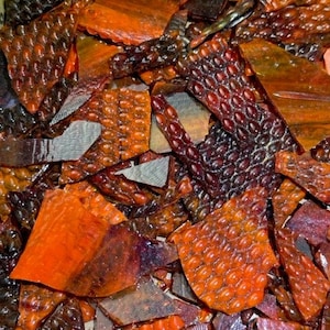 May include: A collection of irregular, broken glass shards in shades of deep amber, burnt orange, and dark brown. The pieces feature a distinct, raised, repetitive oval-shaped texture on one side, while other fragments are smooth and translucent.