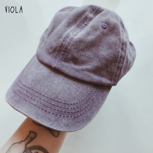 K&ouml;nnte beinhalten: Eine lilafarbene Baseballkappe mit leicht abgenutztem Look. Die Kappe hat einen gebogenen Schirm und ein gesticktes Design.