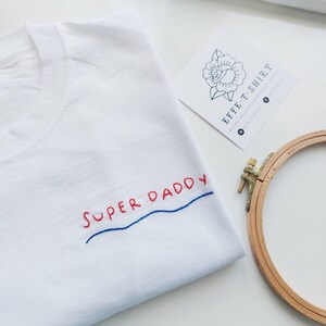 May include: White t-shirt with the phrase "SUPER DADDY" embroidered in red and blue thread. The shirt is folded and lying on a white surface. A wooden embroidery hoop is in the background.