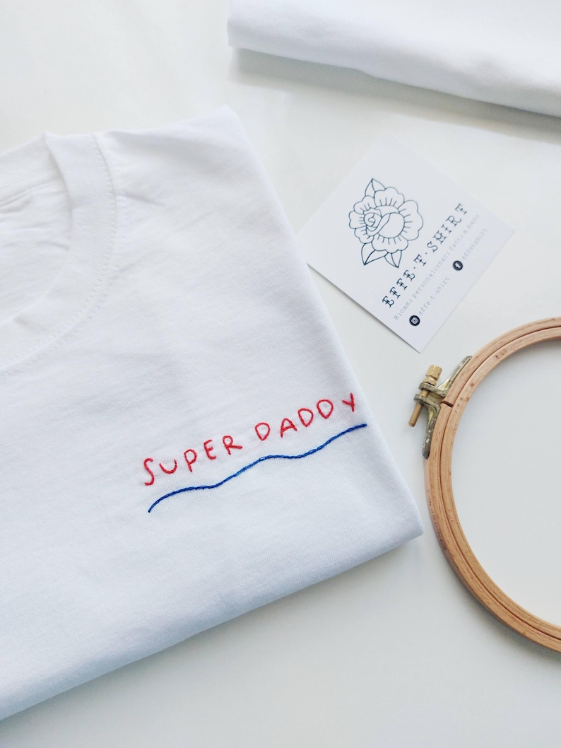 May include: White t-shirt with the phrase "SUPER DADDY" embroidered in red and blue thread. The shirt is folded and lying on a white surface. A wooden embroidery hoop is in the background.