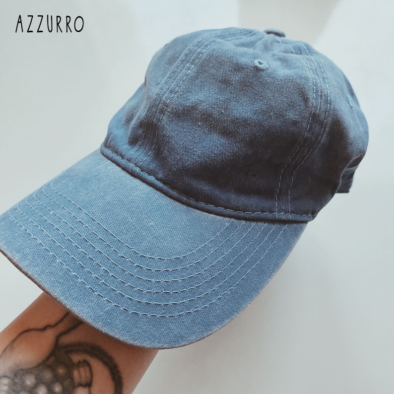 K&ouml;nnte beinhalten: Eine blaue Baseballkappe aus Denim mit einem gebogenen Schirm. Die Kappe hat ein gesticktes Design auf dem Schirm.