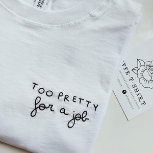Può includere: T-shirt bianca con la frase ricamata "TOO PRETTY for a job" in corsivo nero. È visibile un piccolo biglietto con un disegno floreale e la scritta "EFFE T-SHIRT".