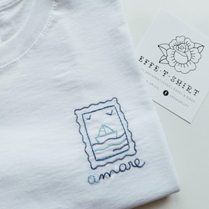 Può includere: T-shirt bianca con un disegno blu ricamato di una barca a vela sull'oceano, incorniciato come un francobollo. La parola "amare" è ricamata sotto il disegno. Una carta con un'illustrazione di rosa e il testo "EFFE.T.SHIRT" è sullo sfondo.
